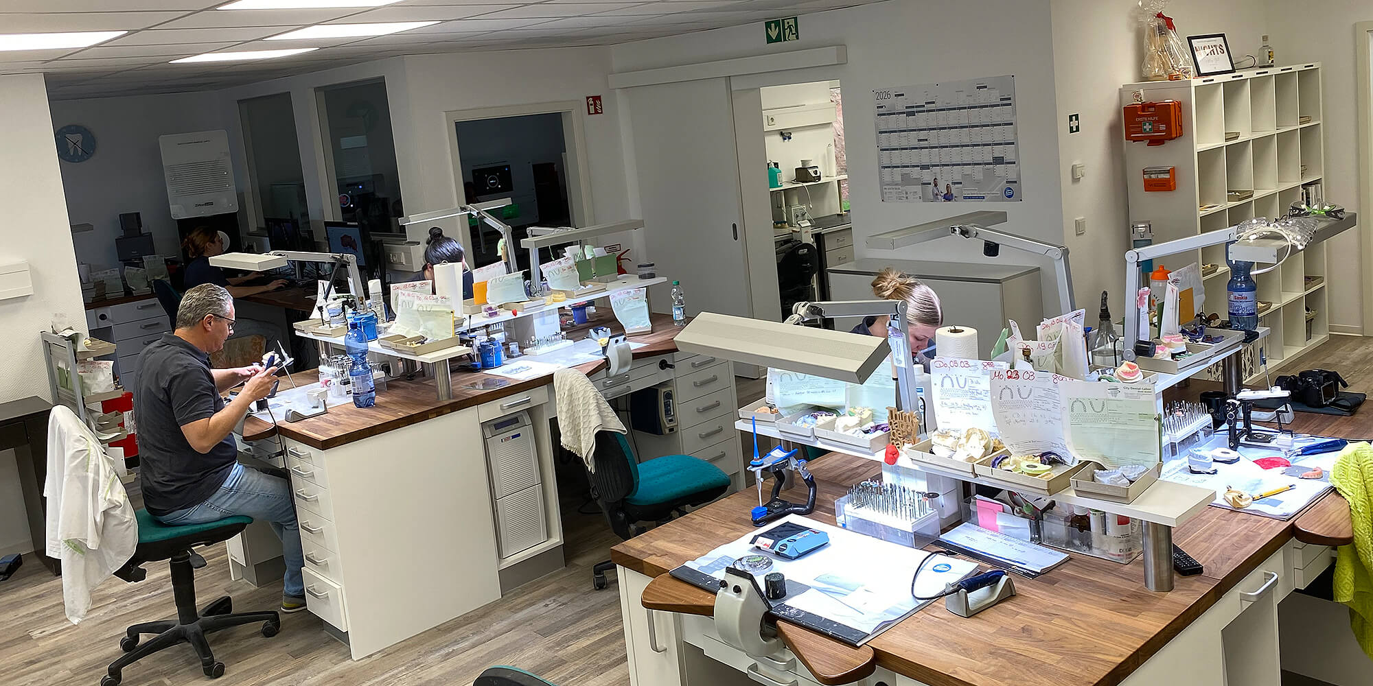 Blick in das helle Dentallabor mit mehreren Arbeitsplätzen, Werkzeugen und Zahntechnikerinnen und Zahntechnikern bei der Arbeit