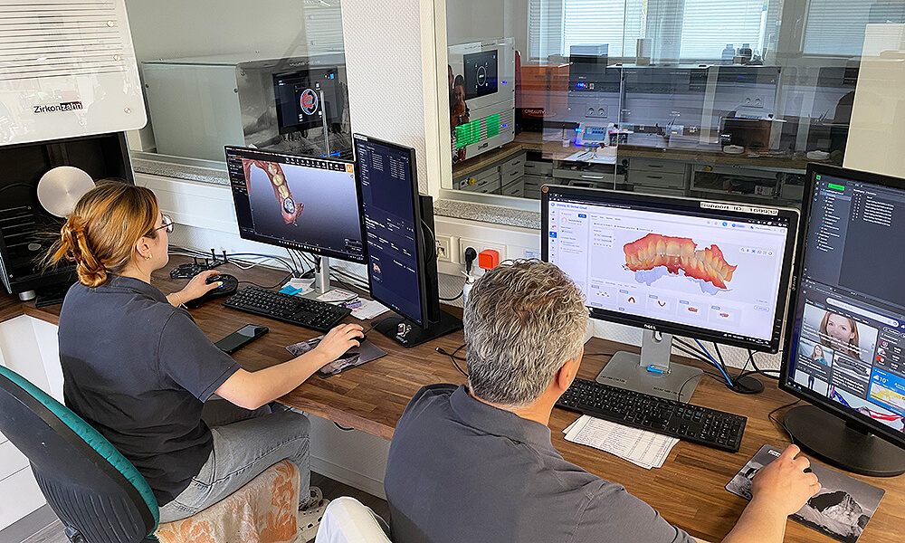 Zwei Zahntechniker bei der digitalen CAD-Konstruktion von Zahnersatz an mehreren Computerarbeitsplätzen im Dentallabor.