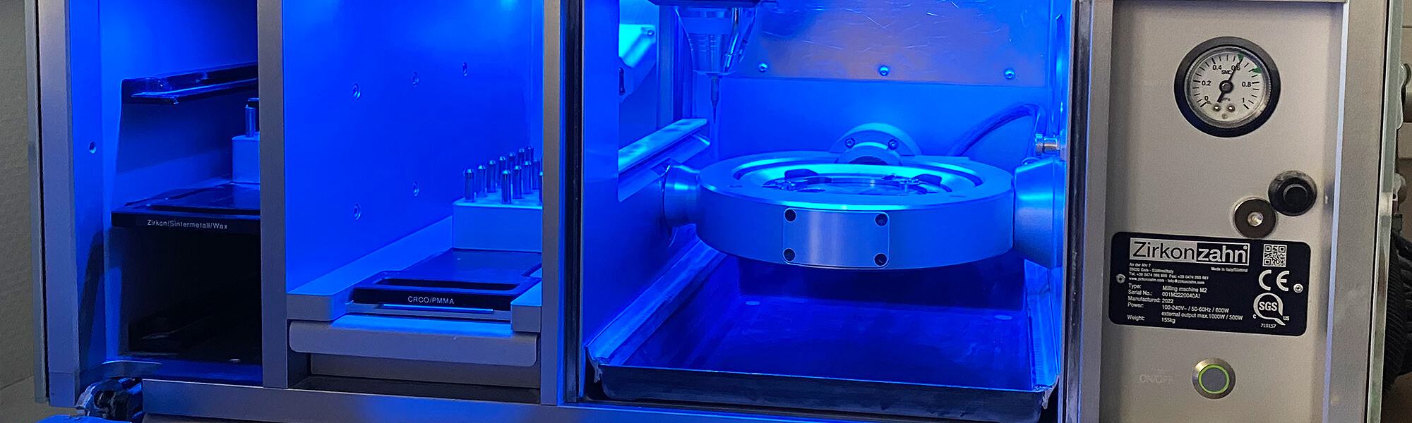 Geöffnete CAD-CAM-Fräsmaschine mit blau beleuchtetem Innenraum und Werkzeugaufnahme für die digitale Zahnersatzfertigung.