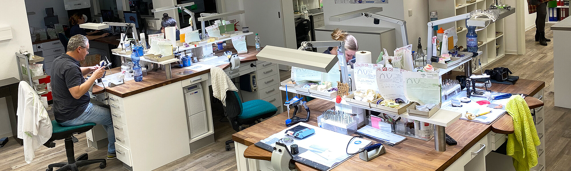 Zahntechnische Arbeitsplätze im Dentallabor mit Modellen, Werkzeugen, Lampen und Mitarbeitern bei der manuellen Bearbeitung.